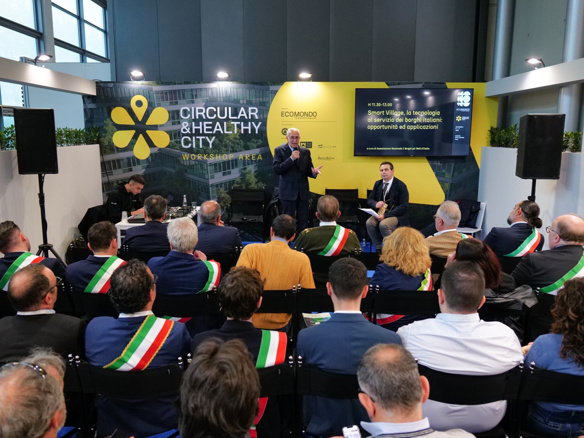 Numerosi sindaci, riconoscibili dalla fascia tricolore, partecipano ad una conferenza con un uomo che parla sul palco accanto ad uno schermo che riporta la scritta 'smart village, la tecnologia al servizio dei borghi italiani: opportunità e applicazioni'