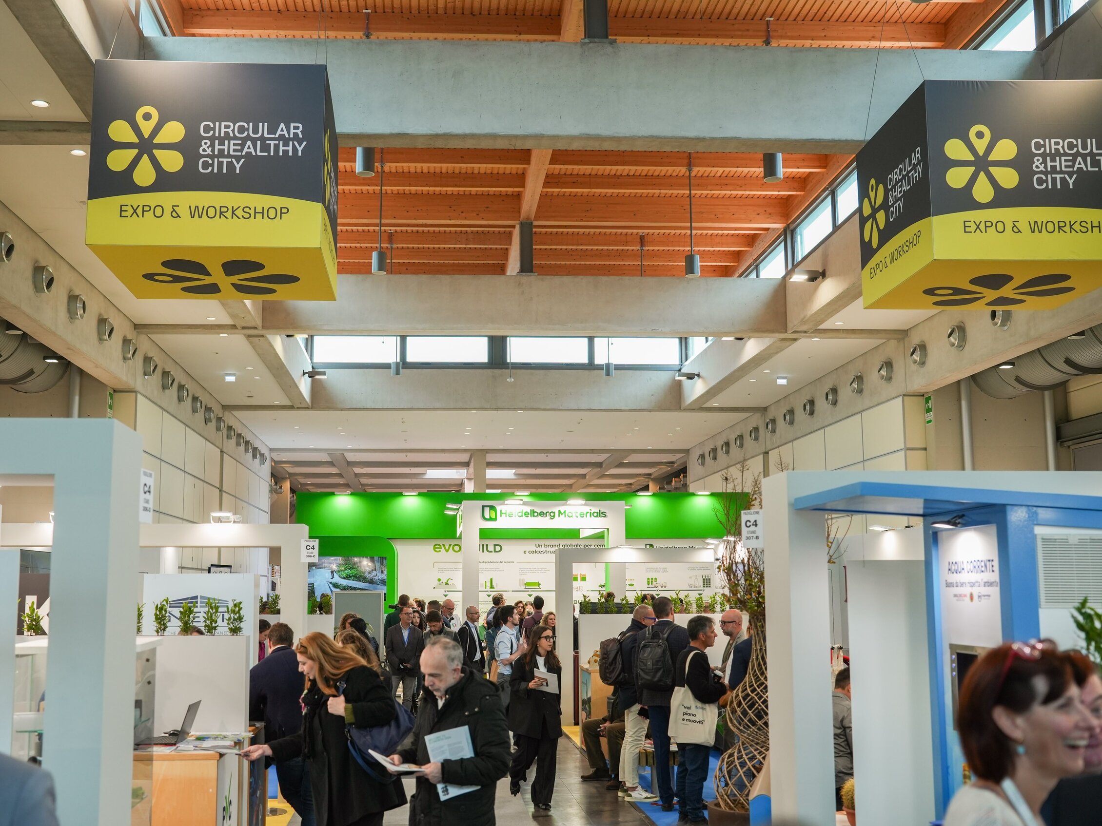 Visitatori di Ecomondo esplorano gli stand sotto grandi pannelli sospesi con la scritta 'Circular & Healthy City – Expo & Workshop'