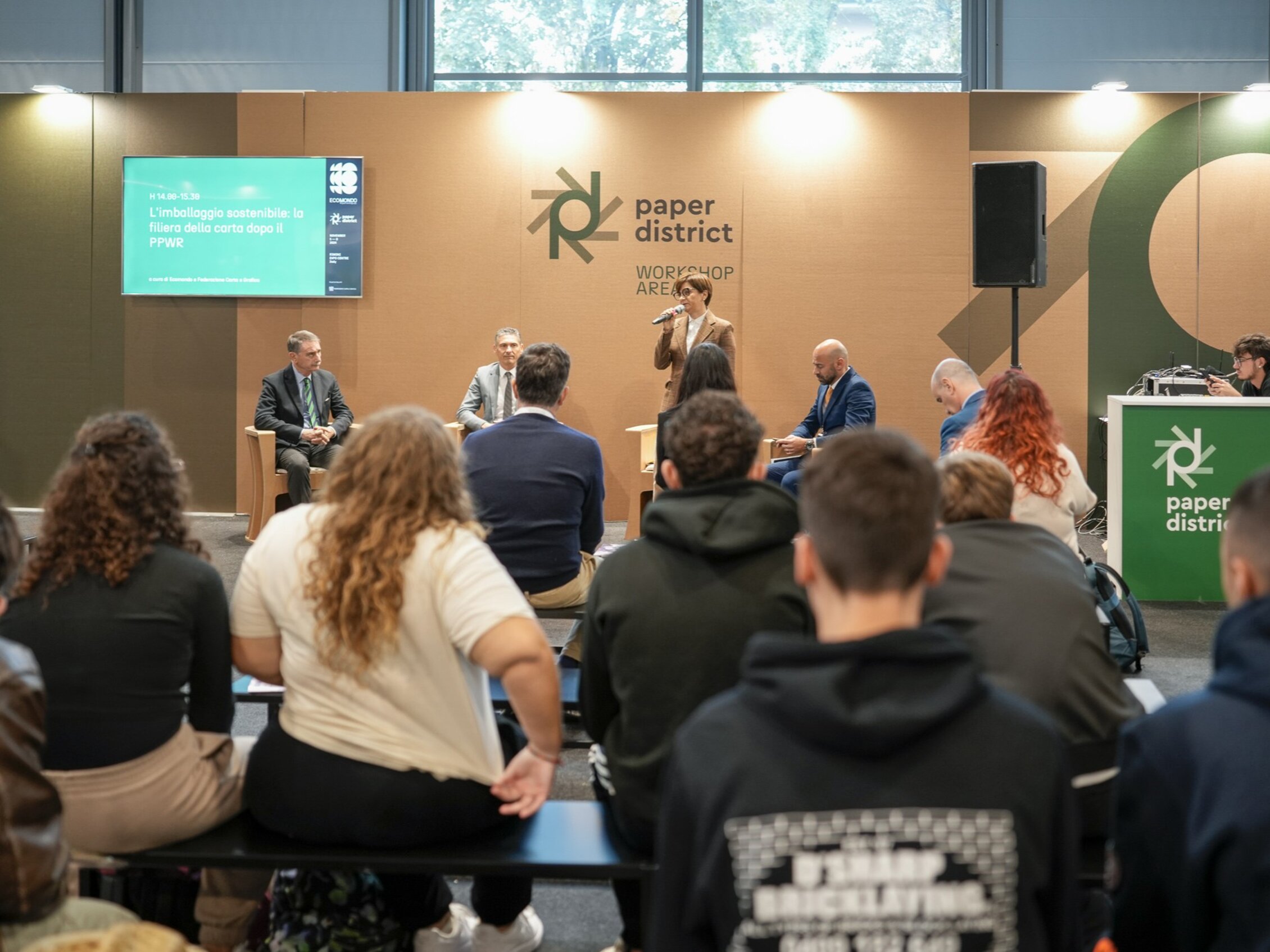 Partecipanti assistono a una conferenza nell'area 'Paper District' dove una donna con il microfono parla su un palco con alle spalle una grande parete di cartone 