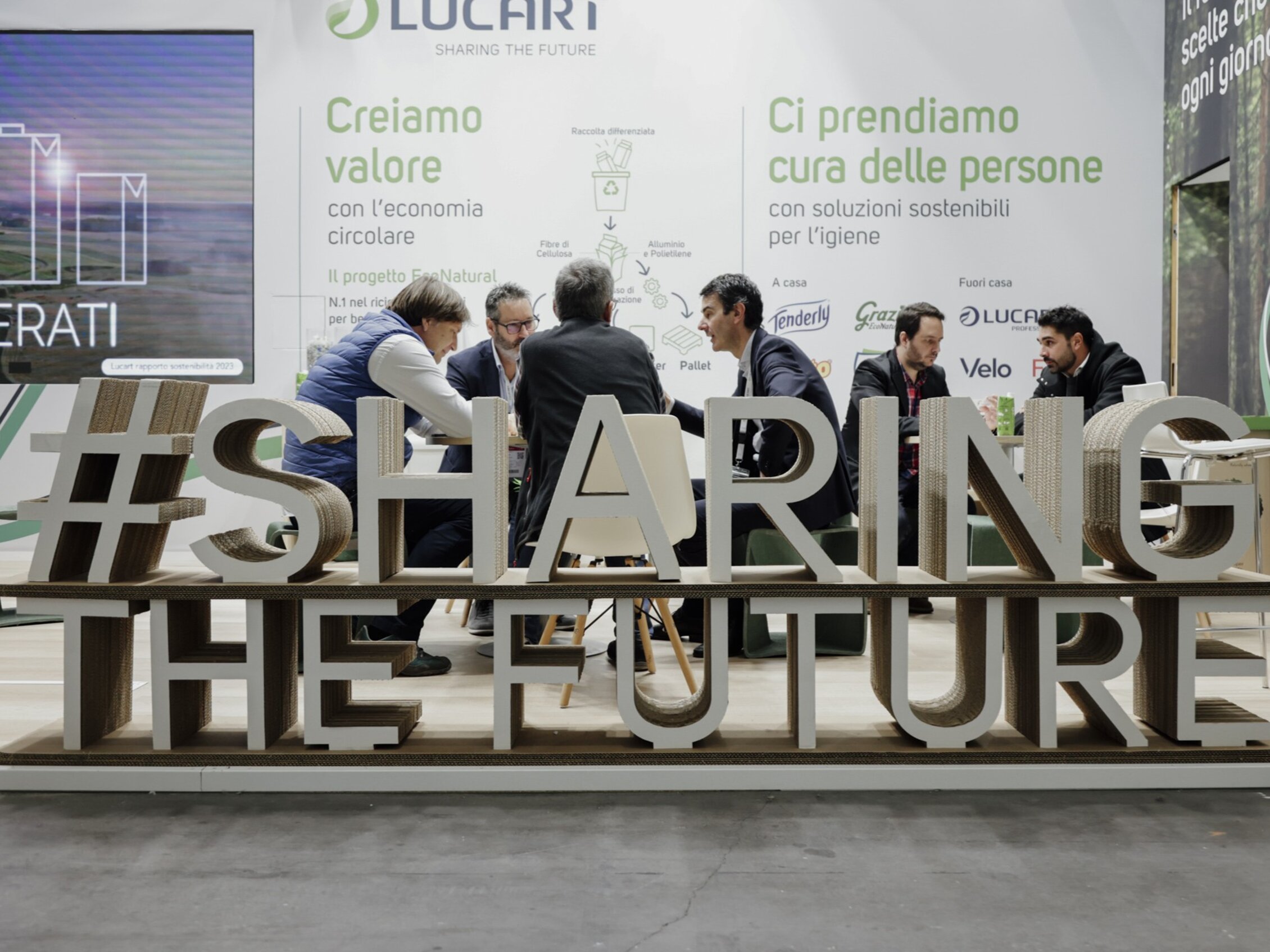 figure professionali parlano seduti a un tavolo davanti a pannelli informativi sull’economia circolare e dietro una grande installazione con la scritta 'SHARING THE FUTURE'