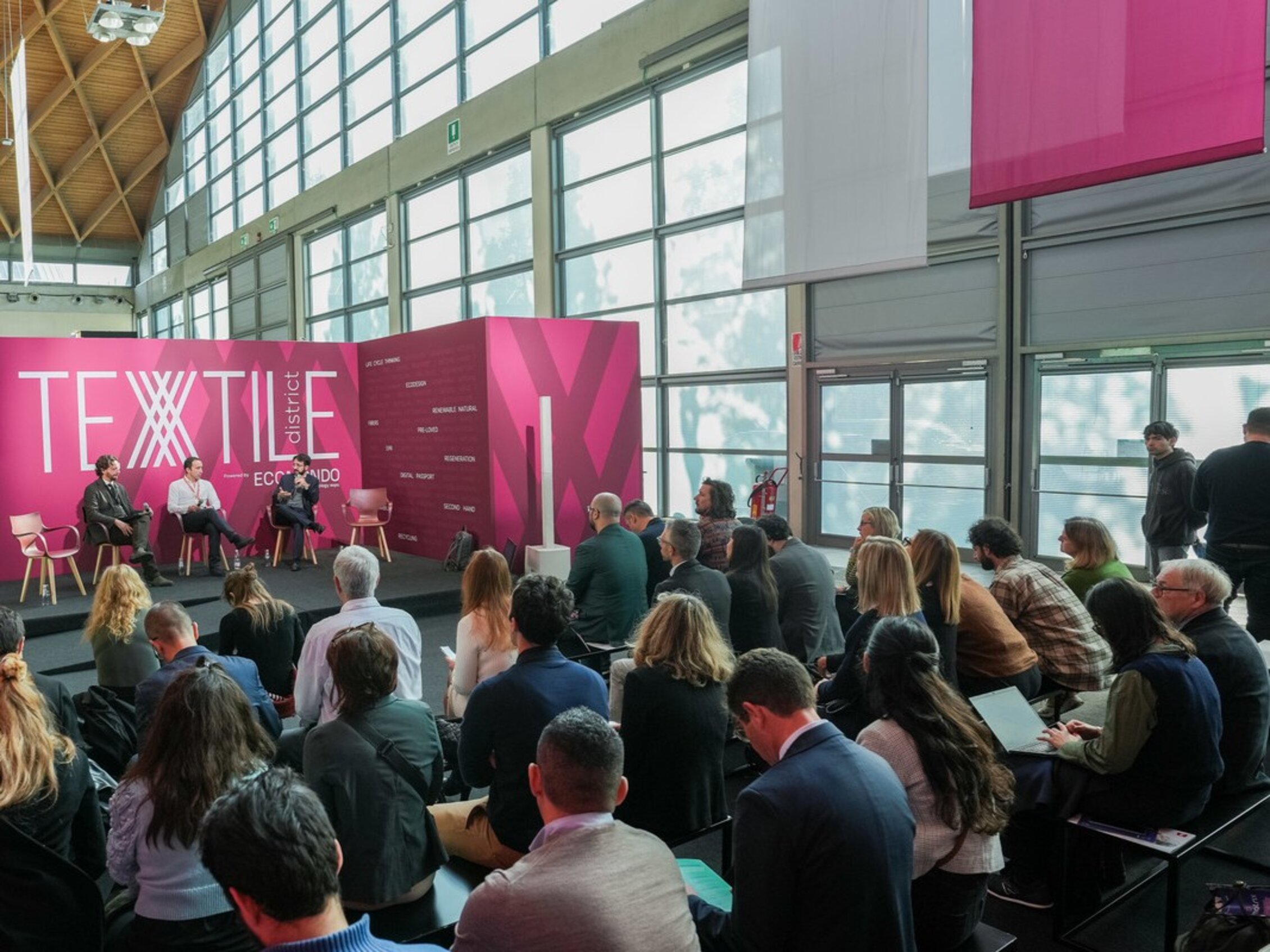 Pubblico assiste a una conferenza nell'area 'Textile District' con tre relatori seduti su un palco decorato in porpora
