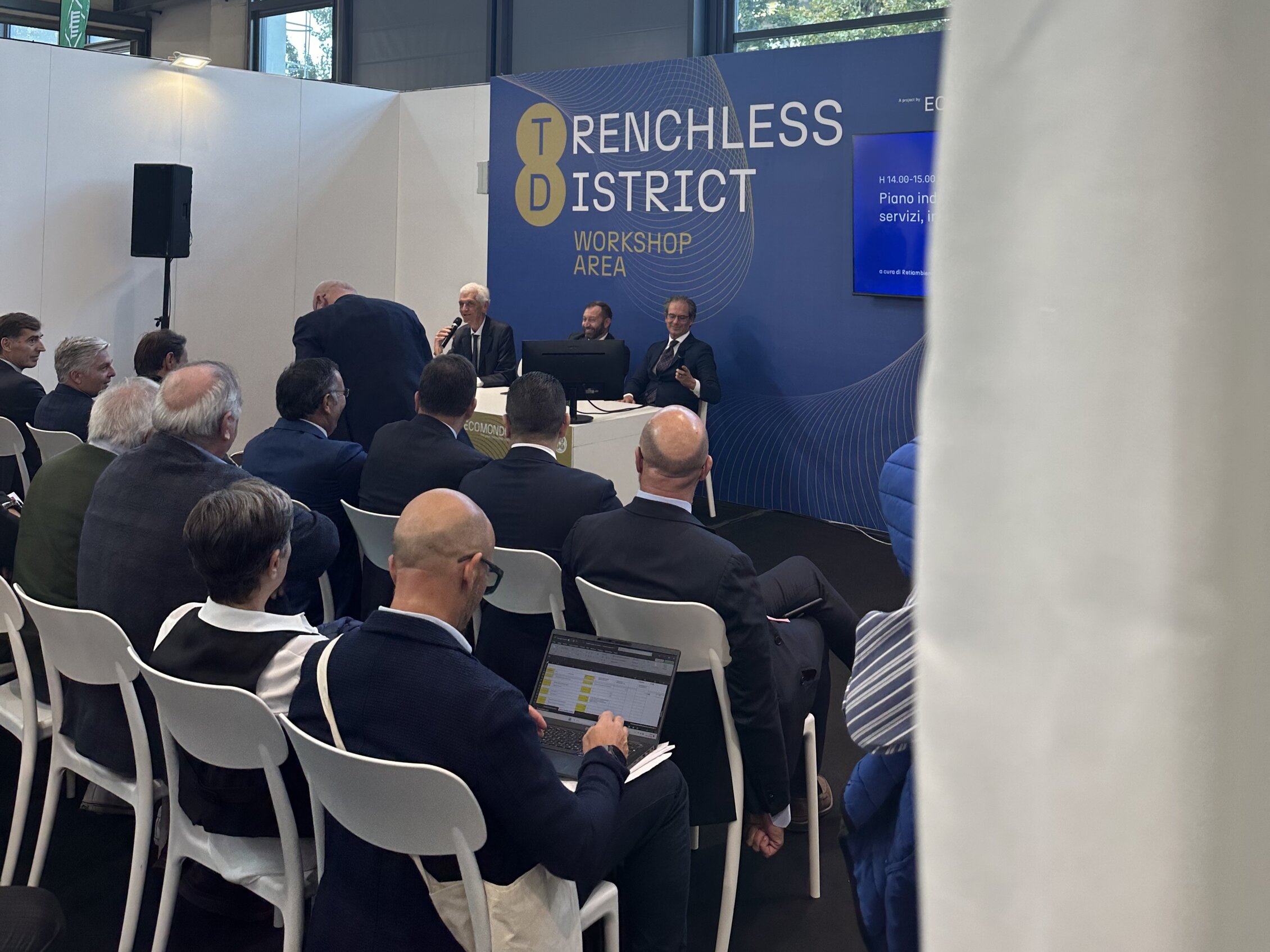Un gruppo di partecipanti assiste a un workshop nella zona 'Trenchless District', dove tre relatori sono seduti su un palco