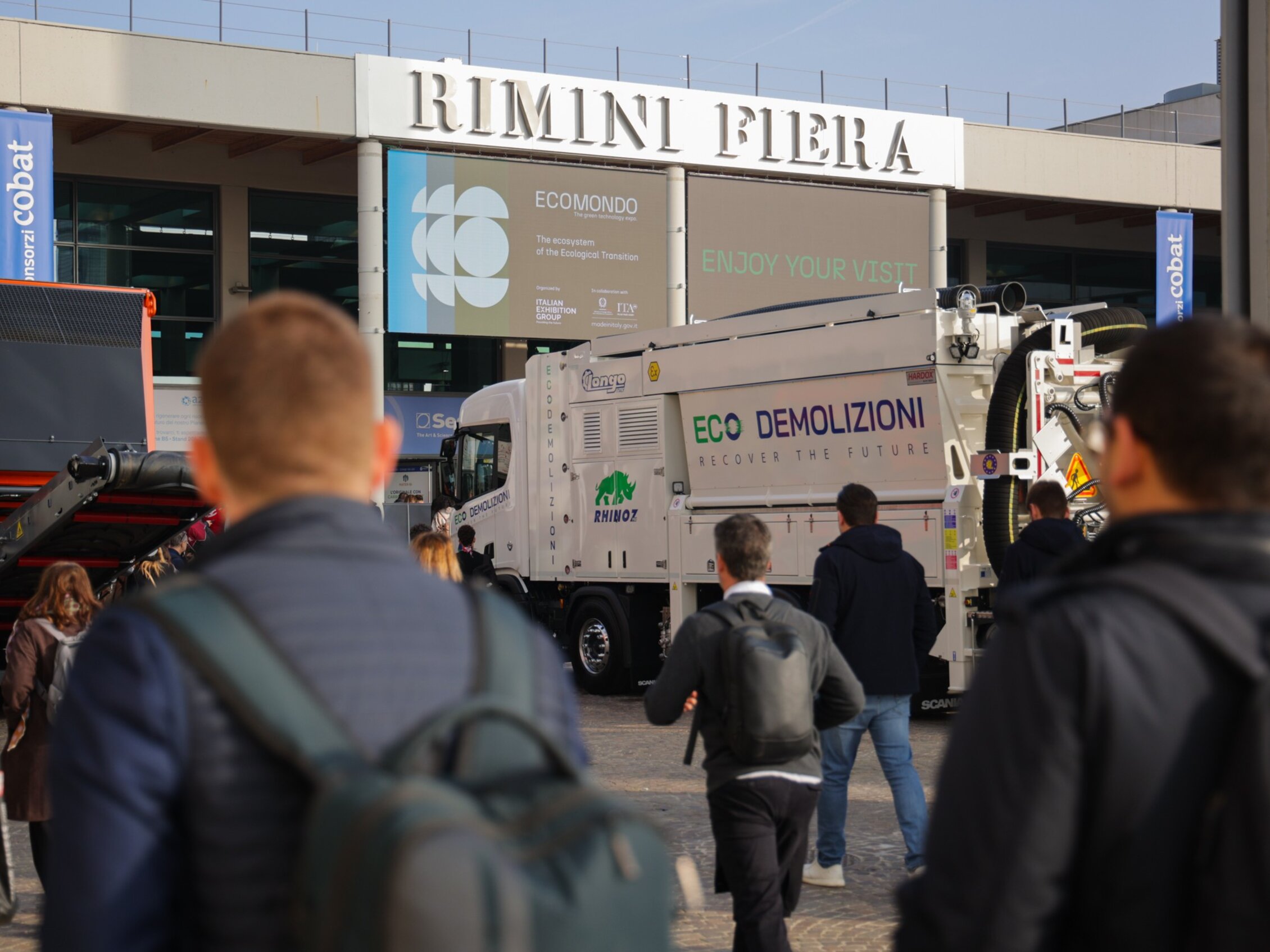 partecipanti ad ecomondo camminano verso l'ingresso della fiera, di fronte un camion bianco con la scritta verde 'ECO DEMOLIZIONI'