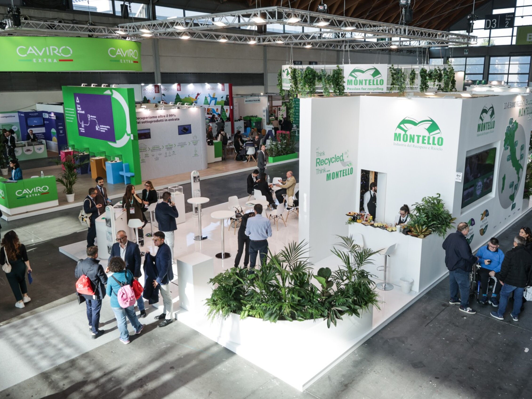 visitatori della fiera circondano gli stand di Caviro extra e Montello che riporta lo slogan 'The Recycled is the New Raw' su ampie superfici bianche, arricchite da elementi verdi e piante ornamentali.