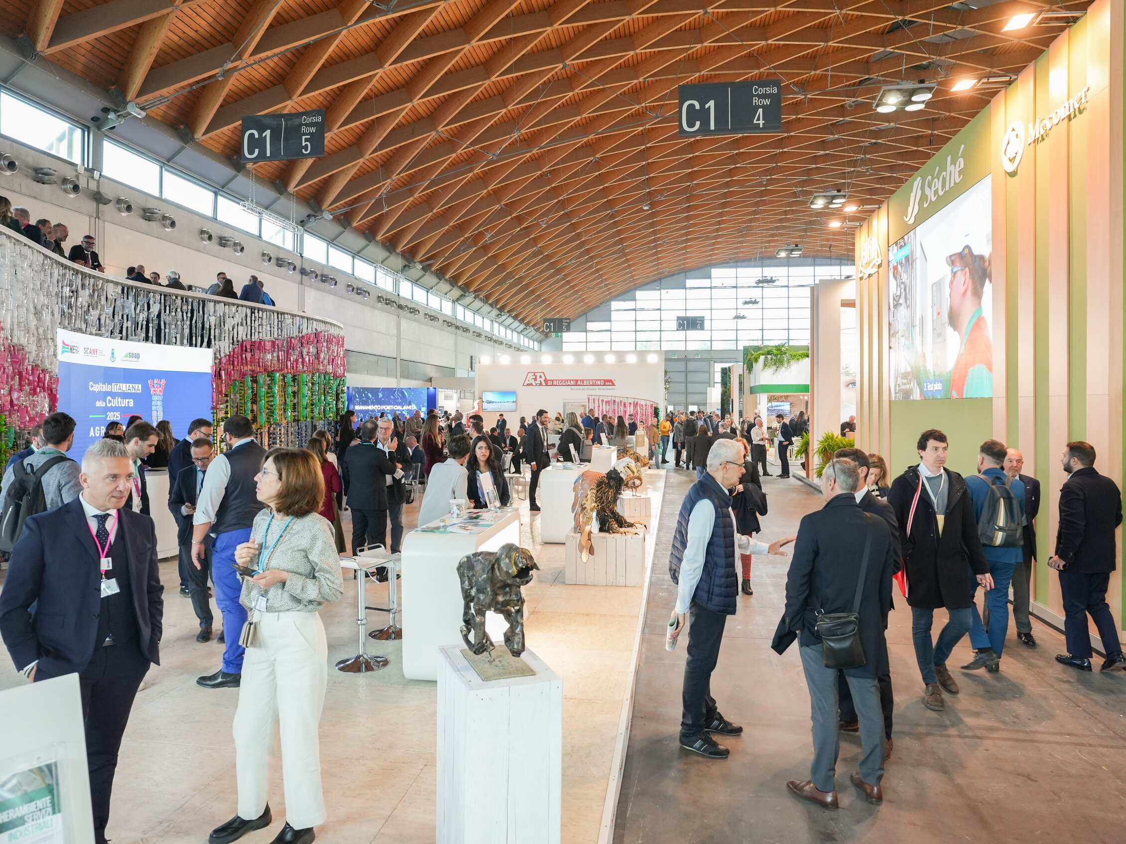 Visitatori esplorano l'area espositiva affollata di Ecomondo, tra stand, installazioni artistiche e decorazioni colorate sotto il soffitto arcuato in legno della Fiera di Rimini
